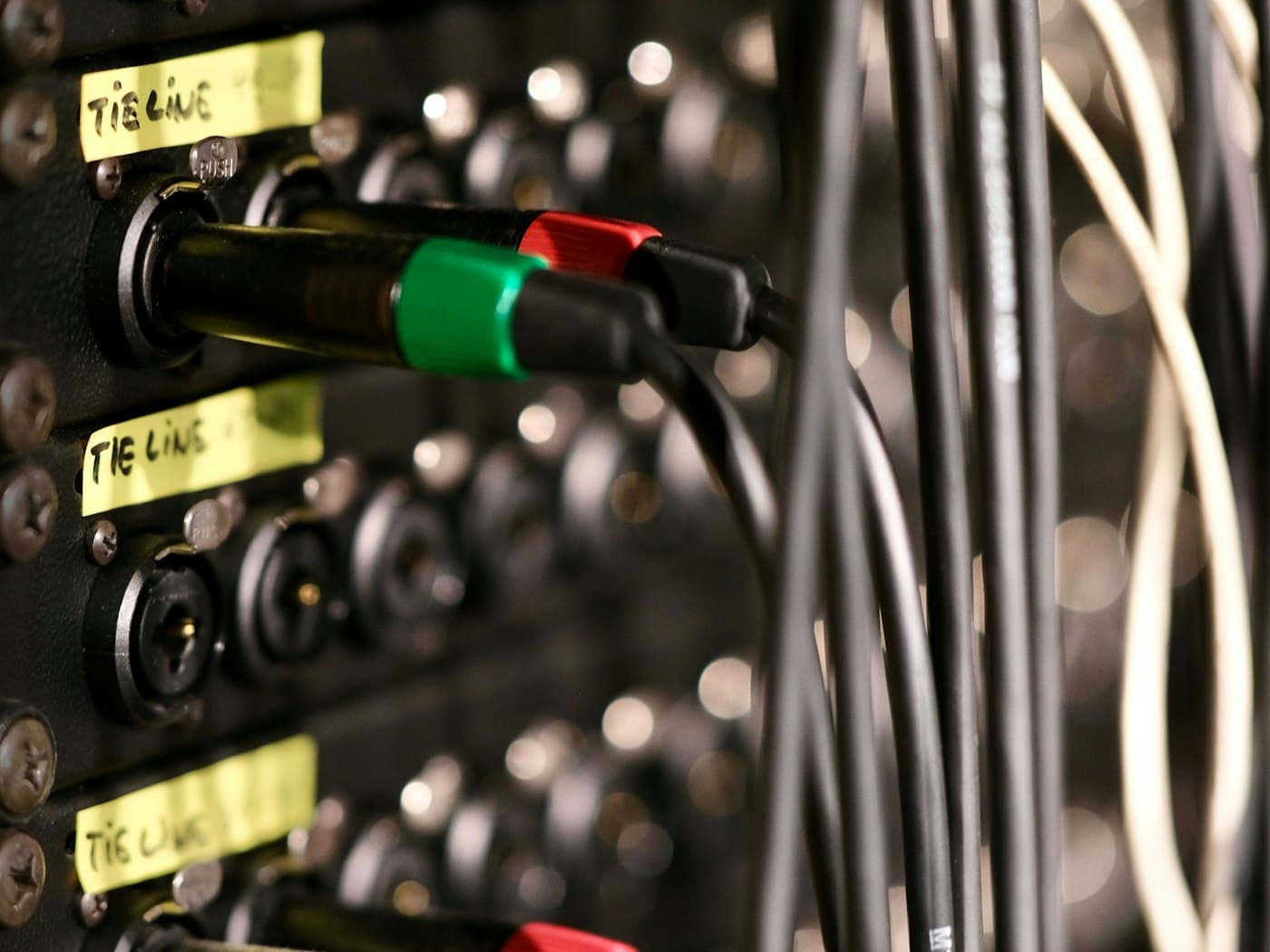 Close-up of audio cables plugged into a rack patch bay with hand-written yellow tie-line labels.