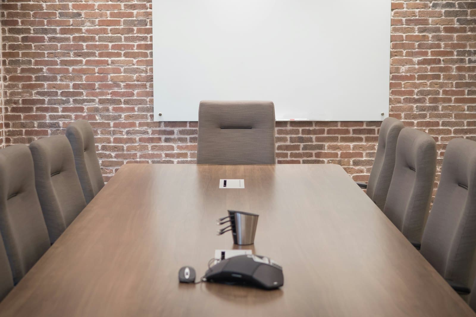 An empty conference room: a long wooden table with a conference phone and cable cubby, upholstered chairs, and a projection screen on a brick wall.