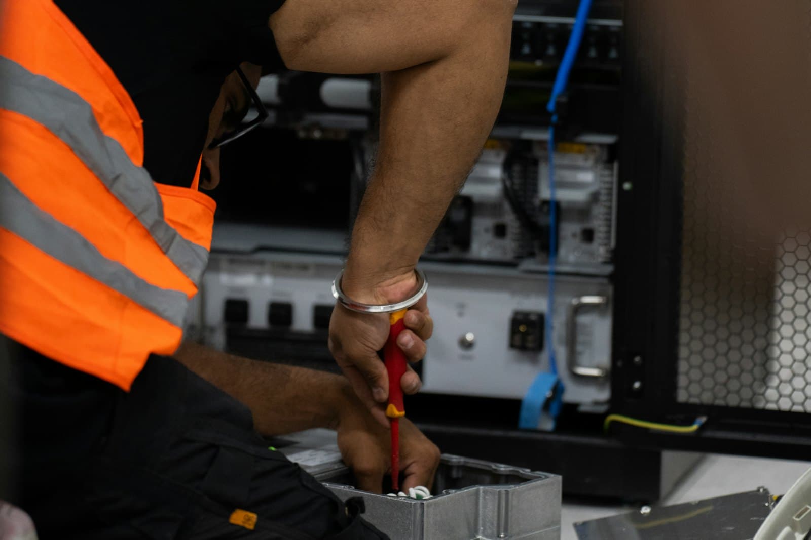 A technician in a high-visibility vest works inside an equipment rack with a screwdriver, cabling and rack-mounted hardware behind.
