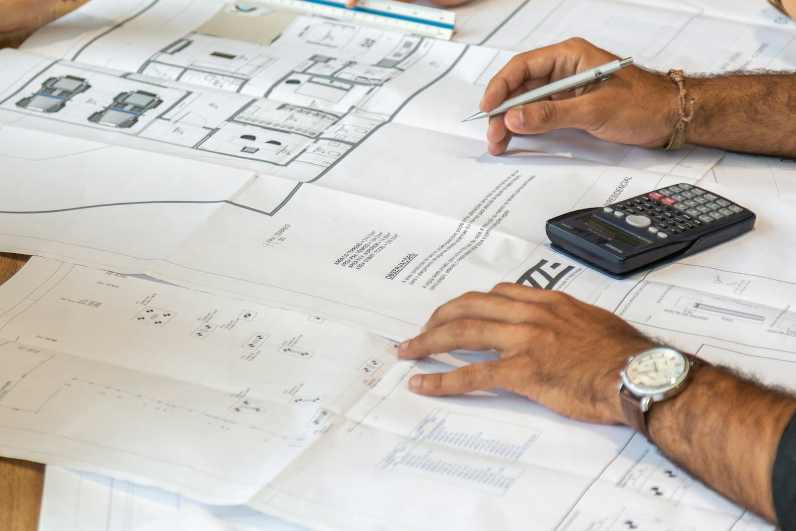 Hands with a pencil reviewing printed building plans on a desk, a calculator beside them.