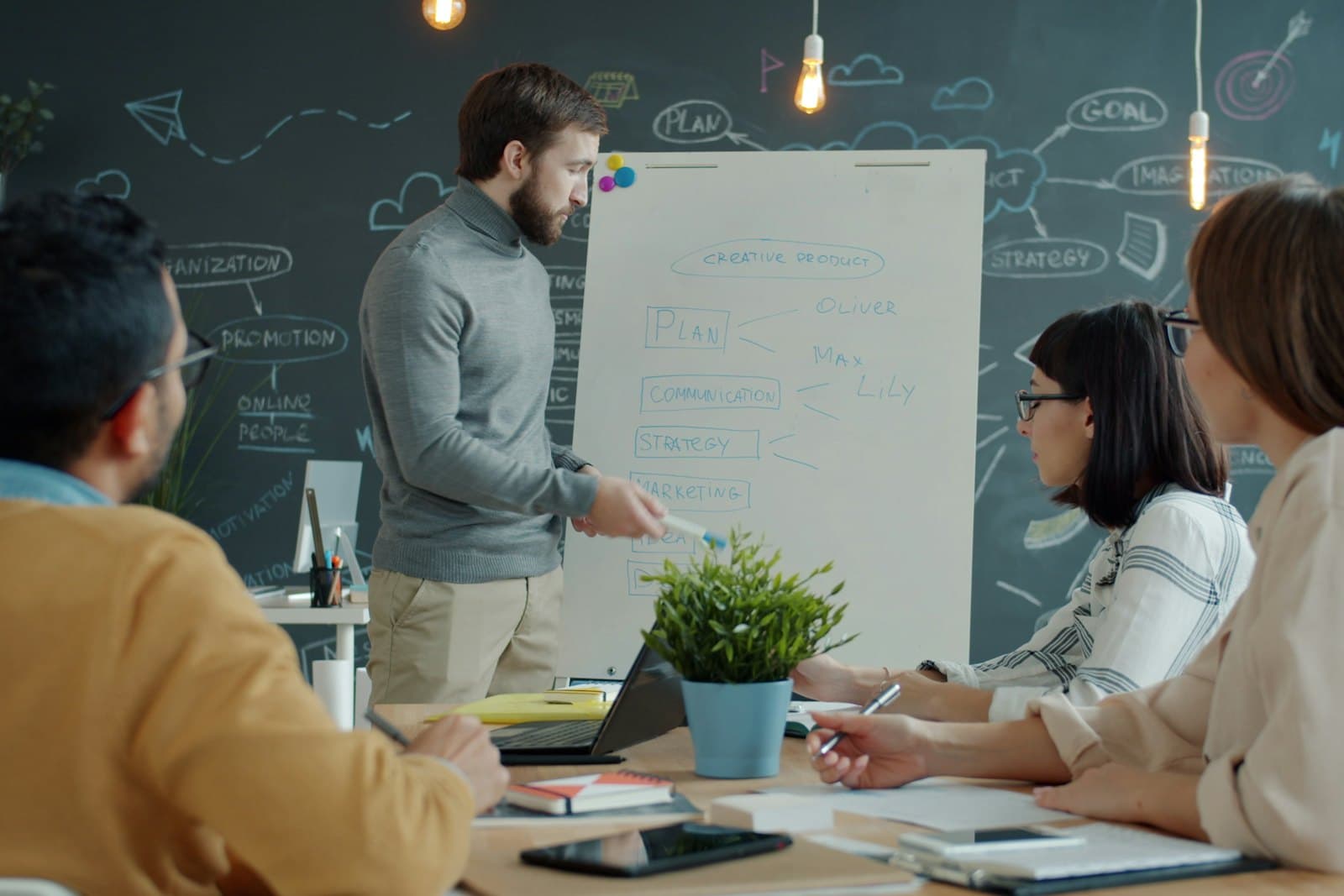 A colleague explains a plan on a whiteboard while three teammates at a table listen and take notes.