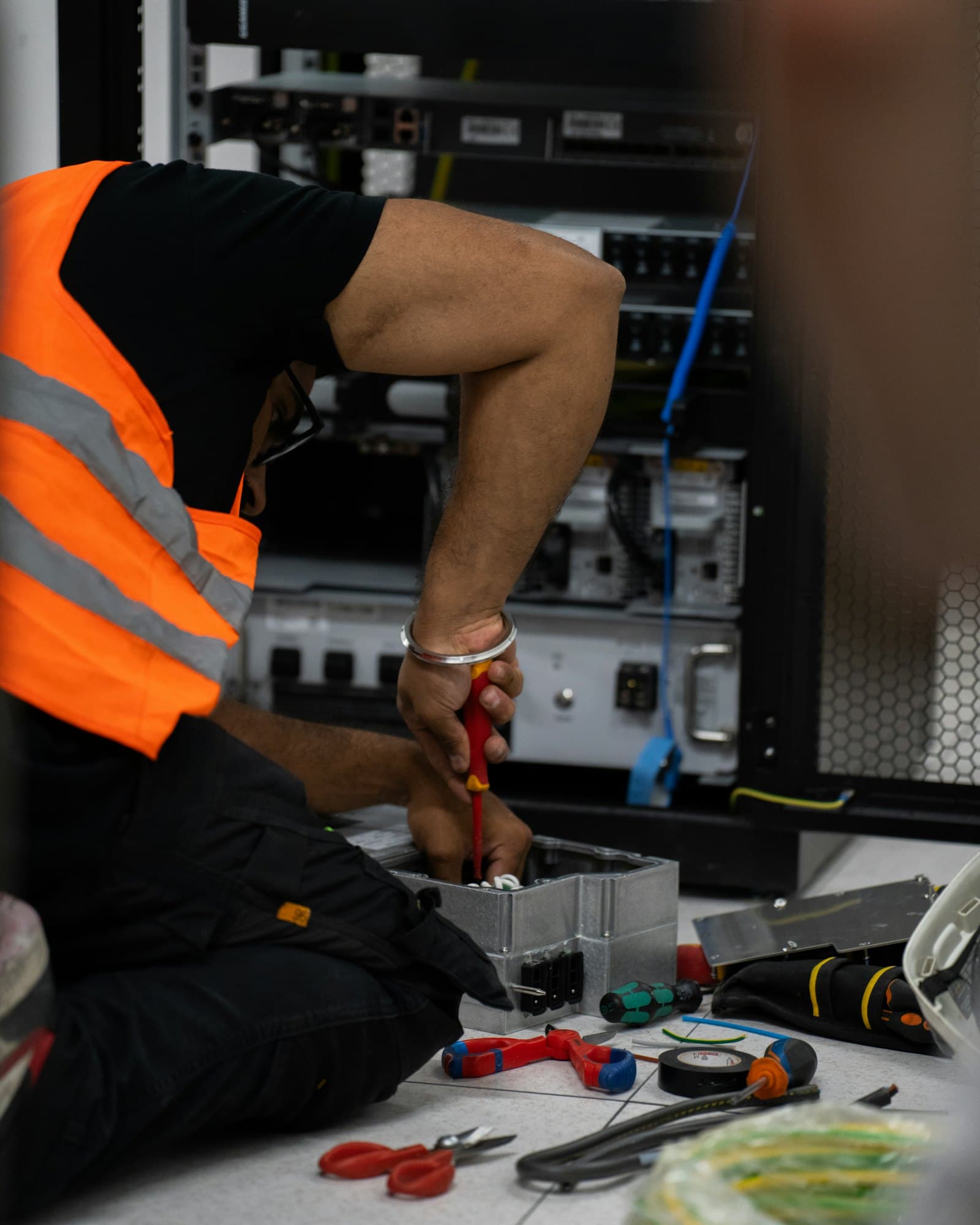 A technician in a high-visibility vest works with a screwdriver in front of an open equipment rack, tools spread on the floor.